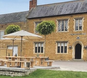 The Nevill Arms — the honey-ironstone facade in sun, mullioned windows, clipped bay trees and an arched door, benches on the forecourt